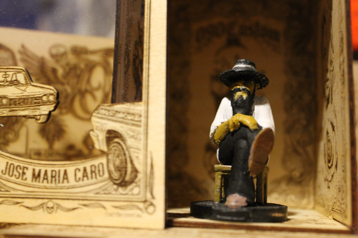 
"Fotografía en primer plano de una figura de colección a escala (5-6 cm) de Don Memo, un hombre mayor con sombrero, barba larga, camisa blanca y pantalones oscuros. Está sentado de perfil sobre una silla en miniatura, con las manos cruzadas en su regazo y la mirada hacia el frente, transmitiendo una postura serena y experimentada. La figura, de estilo artesanal y pintada a mano, se encuentra dentro de un nicho de madera grabado."