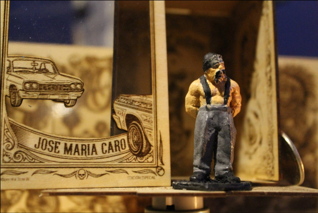 Oso
"Fotografía en primer plano de una figura de colección a escala (5-6 cm) de Oso, un hombre de físico musculoso con una camiseta de tirantes negra y pantalones claros. Lleva una pañoleta tipo bandana de color oscuro en la cabeza. Está de pie en posición desafiante, con los brazos cruzados en el pecho, mostrando una expresión de respeto y autoridad. La figura está pintada a mano y se exhibe en un nicho de madera con detalles grabados."