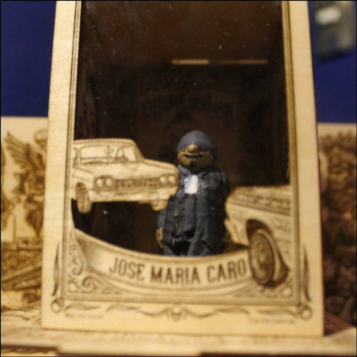 Silencio
"Fotografía en primer plano de una figura de colección a escala (5-6 cm) de Silencio, un personaje corpulento vestido con una campera de jeans gris y un gorro pasamontañas del mismo color que cubre su cabeza y parte de su rostro. Está de pie en posición firme, de frente a la cámara, con los brazos a los lados. La figura presenta un acabado artesanal y está colocada dentro de un compartimento de madera con grabados decorativos."
