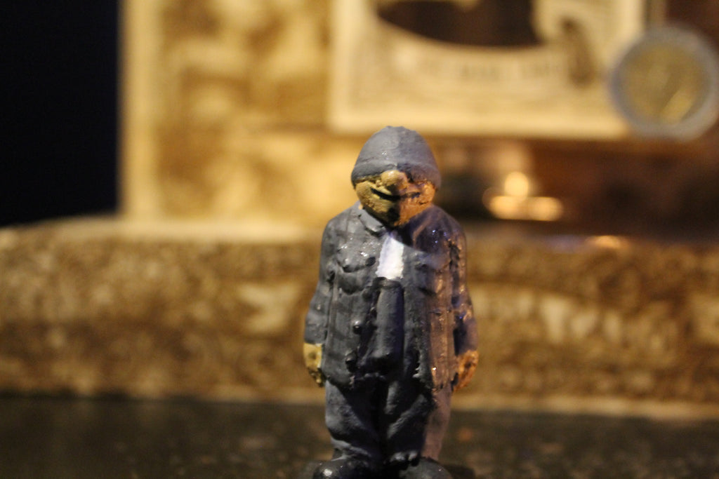 Silencio
"Fotografía en primer plano de una figura de colección a escala (5-6 cm) de Silencio, un personaje corpulento vestido con una campera de jeans gris y un gorro pasamontañas del mismo color que cubre su cabeza y parte de su rostro. Está de pie en posición firme, de frente a la cámara, con los brazos a los lados. La figura presenta un acabado artesanal y está colocada dentro de un compartimento de madera con grabados decorativos."